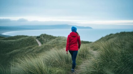 Female adult walking on a winding path through tall grassy dunes. She explores the vast coastal landscape under a cloudy sky. A sense of adventure and freedom in nature