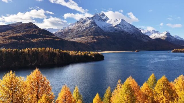 A peaceful autumn view of Lake Sils (Silsersee) in Switzerland&rsquo;s Engadin valley. Golden larch forests meet calm blue waters, with small residential houses resting along the alpine edge.