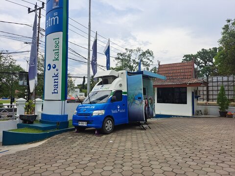 A blue and white Bank Kalsel mobile ATM service vehicle (Layanam Mobile ATM) parked outdoors next to an ATM sign. The vehicle's side is open, showing a service window and steps