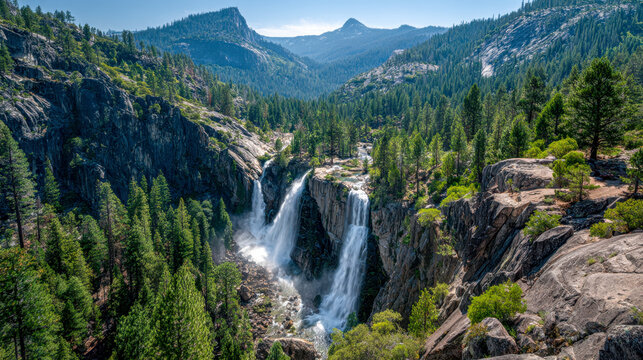 Majestic twin waterfalls cascading through a lush green forest with rugged mountain peaks and rocky cliffs under a clear blue sky in a scenic wilderness landscape