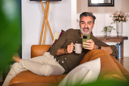 Man relaxing on sofa at home with coffee and using phone
