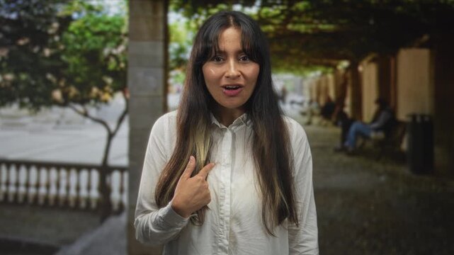 Woman points finger to shirt on street under stone arcade, eyes closed with gentle smile; self affirmation confidence.