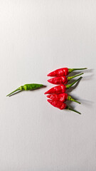 A single green chili stands apart from a cluster of red chilies on a clean white surface, creating...