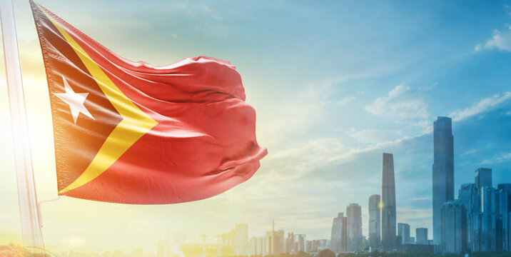 East Timor flag wavering in beautiful clouds with building skyline. The flag with drone shot in beautiful sky. East Timor national flag for independence day.