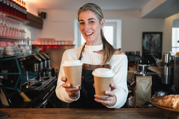 A friendly blonde waitress offers a to go coffee while the cafe owner stands behind the counter showing strong teamwork and service