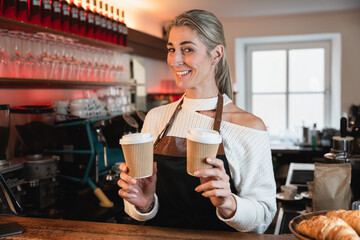 A smiling blonde waitress presents takeaway coffee with the cafe owner working behind the counter showing strong service values