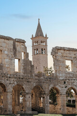 Obraz premium Ancient stone arches and weathered walls with a tall historic bell tower rising in the background. Warm sunlight highlights the detailed masonry and classic architectural forms.