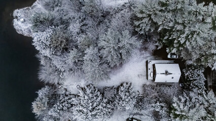 Aerial view of a snow-laden cabin embraced by frosted trees, on the shore of dark waters of a tranquil lake. White Mountains, New Hampshire, USA.
