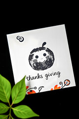 A minimalist black and white Thanksgiving card on a dark surface, featuring a cute, textured pumpkin stamp design and decorative doodles, accented by a single bright green leaf.