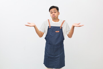 Confused and upset young Asian man in apron shrugs his shoulders with his arms out to the side, feeling frustrated, unable to understand what is happening, standing on a white background.