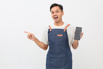 Young Asian man, barista, barman, wearing blue apron, showing blank area of ​​phone screen, pointing to the side, isolated on white background.