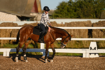 Rider on horse practices basic dressage exercises in open arena near stable, sitting in saddle and wearing helmet. Racing, horse care, and training © GRON777