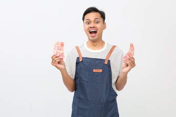 Successful Asian male barista wearing apron feeling relieved and happy, holding Indonesian rupiah banknotes in both hands, smiling looking at camera, isolated on white background.