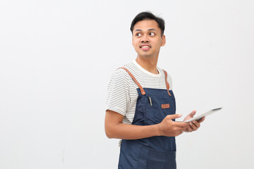 Smiling young Asian male barista wearing blue apron, working in coffee shop or restaurant, holding digital tablet, looking sideways at empty space, isolated on white background.