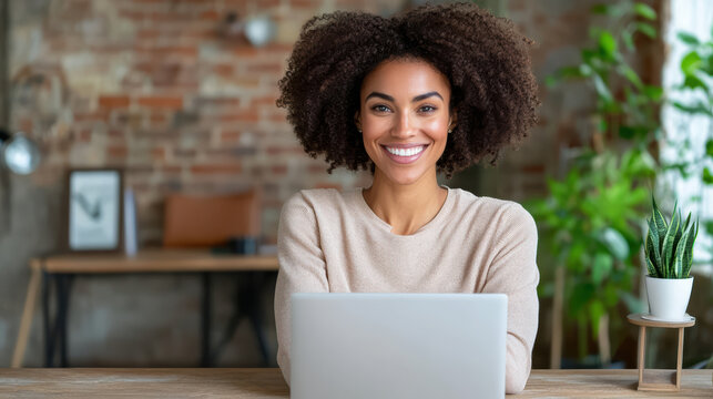 Young woman smiling at laptop in cozy home office with brick wall and plant