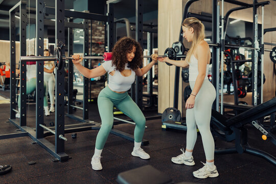 Two women are training together in a gym, with one performing a barbell squat while the other acts as a spotter