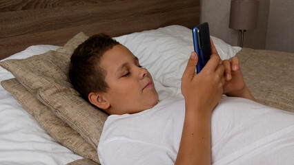 Teenage boy lying in bed, looking at smartphone screen and smiling. Children and gadgets. Communication using a mobile phone. Communication in messengers and mobile applications of gadgets.