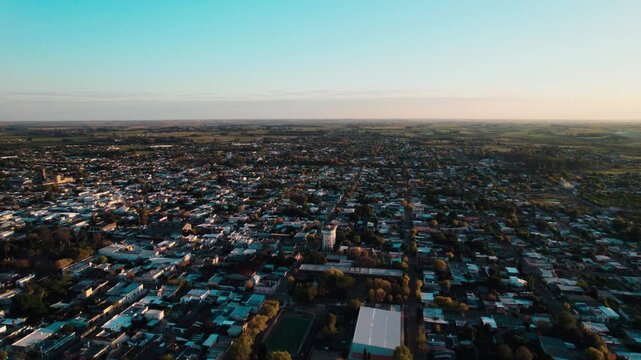 Drone shot capturing the full breadth of Carmelo, Uruguay, with low-rise buildings and tree canopy extending into the horizon under a golden hour sky. Features a peaceful, wide view of the town