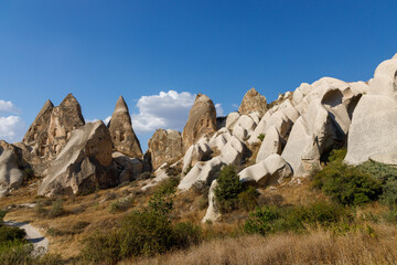 Rock formations in Cappadocia, Turkey, under a clear blue sky. Unique geological shapes with scattered greenery and dry grass create a striking natural landscape.