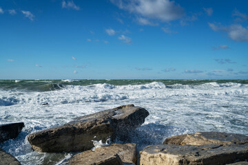 Coastal seascape with large flat rocks in the foreground and turbulent sea waves crashing onto the shore. The sky is clear with scattered clouds. Bright sunlight highlights the texture of wet stone.