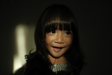 Expressive portrait of a smiling young girl in dramatic shadow lighting. The focused beam of light highlights her joyful expression, creating a warm and cinematic feel with strong contrast.