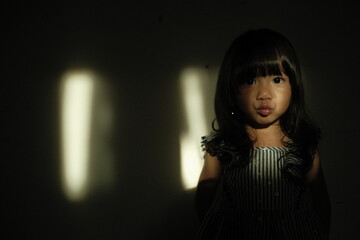 Portrait of a young girl standing against a dark background with cinematic light streaks on the wall. She makes a pout expression, creating a dramatic and artistic mood.