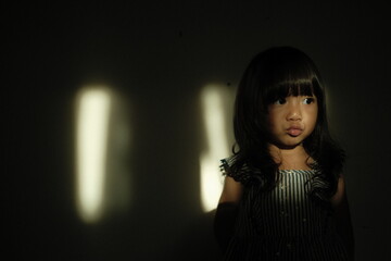Portrait of a young girl standing against a dark background with cinematic light streaks on the wall. She makes a pout expression, creating a dramatic and artistic mood.