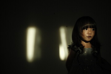 Portrait of a young girl standing against a dark background with cinematic light streaks on the wall. She makes a pout expression, creating a dramatic and artistic mood.