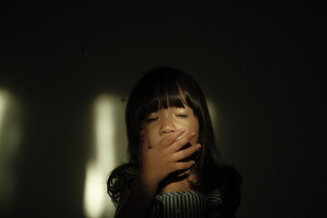 A candid moment of a young girl yawning with her hand covering her mouth while standing in warm dramatic window light. Her eyes are softly closed, creating a natural and intimate expression