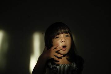 A young girl yawns widely while standing in dramatic window light, her eyes half-open and her hand touching her cheek.