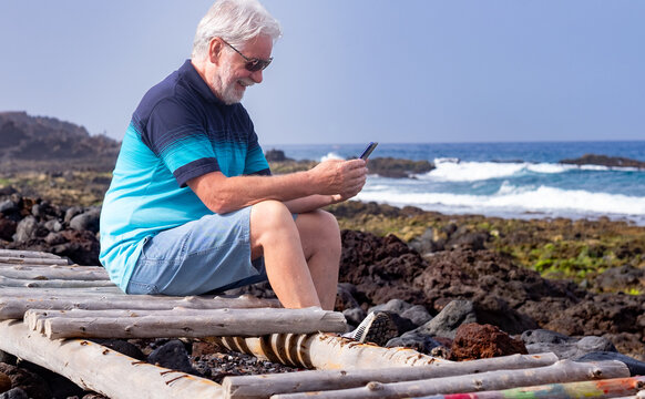 Smiling white haired senior man outdoors face the sea using mobile phone. Relaxed elderly man sitting on rocky beach enjoying tech and social, retirement concept - Powered by Adobe