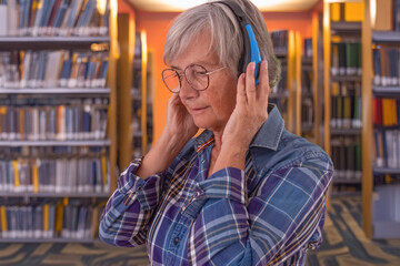 Senior woman with headphones standing in a library, listening to text or online lesson. Concept of lifelong learning, education, technology, and active aging