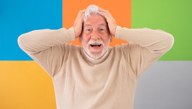 Smiling bearded senior man standing in front of a colorful geometric background, touching his head looking at camera. Concept of decision-making, ideas, creativity, and contemplation