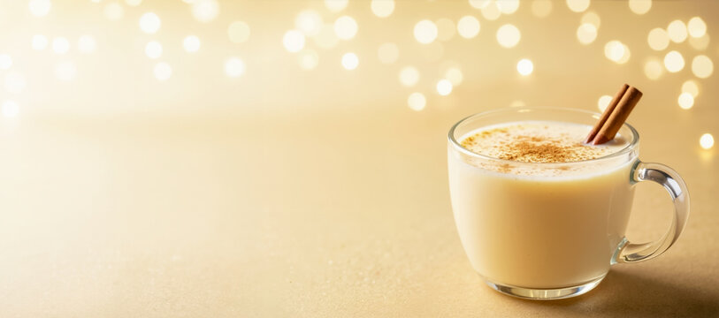 Traditional Christmas eggnog in a glass mug with a cinnamon stick. Festive holiday winter drink on a golden background with bokeh. Seasonal celebration banner concept - Powered by Adobe