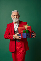 Senior man in a vibrant red suit joyfully carries holiday gifts against a green backdrop