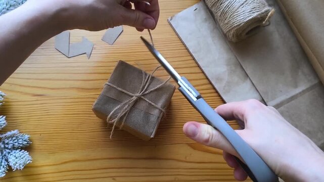 Gift wrapping in eco-friendly materials against a background of a recycling symbol and Christmas tree branches. Scissors trim the ends of a bow.