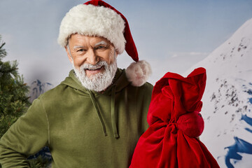 Senior man in winter attire joyfully holding a red sack with a snowy mountain backdrop