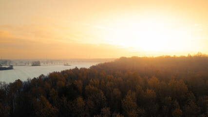 Atmospheric aerial view at sunrise with warm golden light illuminating an autumn forest and a river in the background. The morning fog and the glowing horizon create a calm, peaceful mood in this scen