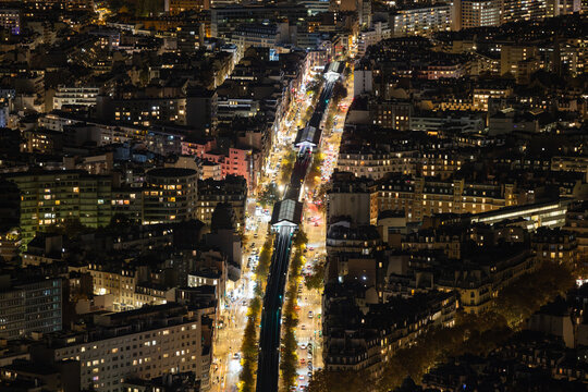Aerial view of a brightly lit railway line cutting through the urban landscape, a glowing artery amidst the dark textures of the city, Paris, Ile-de-France, France.