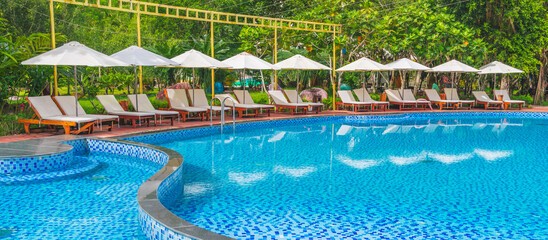 View of the tropical outdoor pool with sun loungers, surrounded by palm trees under a bright sunny sky.