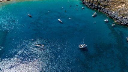 Aerial view of boats scattered across the turquoise waters next to craggy cliffs under the sun, Lindos, Rhodes, Greece.
