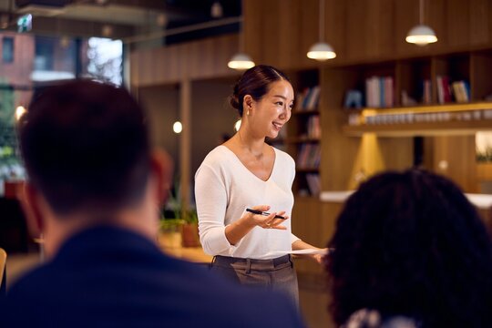 Asian Businesswoman Presenting In Modern Office Meeting With Colleagues And Bookcase Background