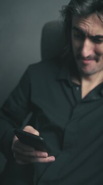 Nodding, comical, agreeable young white European man Slavic handsome expressive, mustached with long hair and large eyes, relaxed and smiling while absorbed in his smartphone, enjoying social media