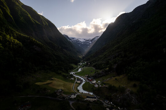 Aerial view of valley with dark, steep mountainsides descending to the river and houses below, with Buarbreen glacier far in the distance, Odda, Vestland, Norway.