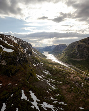 Aerial view of a serene lake nestled between rugged mountains, patches of snow clinging to the slopes under a dramatic sky, Dalen, Telemark, Norway.