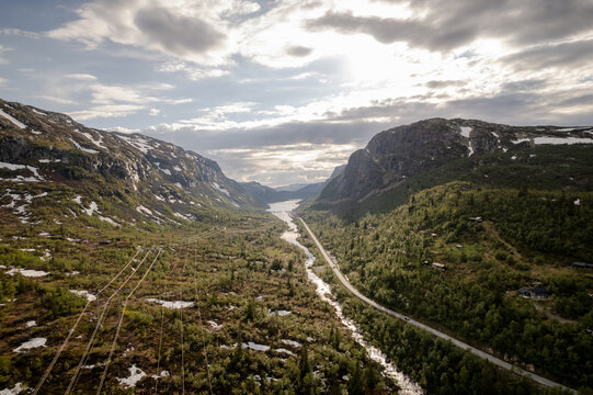 Aerial view of a winding road carving through the lush valley, contrasting with the rugged mountains under a sky, Dalen, Telemark, Norway.