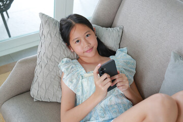 Portrait of lovely young Asian girl relaxing on comfortable sofa, holding a smartphone and looking camera against white background.