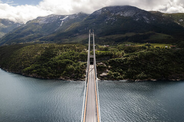 Aerial view of Hardanger Bridge's slender form stretches across the serene Hardangerfjord, connecting verdant shores beneath a crown of misty mountains, Vallavik, Vestland, Norway.
