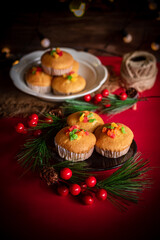 A warm and festive still life of Christmas-themed cupcakes topped with colorful candied fruit. The cupcakes are arranged with holiday decorations including pine branches, red berries, and rustic.