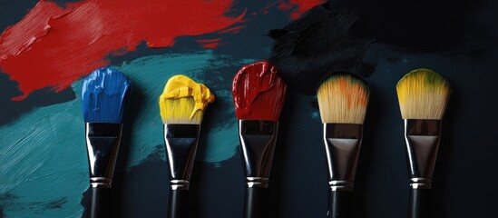 Vibrant paintbrushes with red, blue, yellow, and mixed colors on a dark textured art canvas, overhead view.